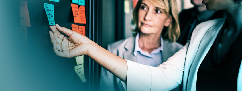 Agiles Projektmanagement: Kolleginnen diskutieren vor einer Ideenwand mit farbigen Post-its, symbolisiert Zusammenarbeit und visuelle Planung in agilen Projekten.