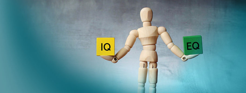 Emotionale Intelligenz: Stilisierte Holzpuppe hält gelben „IQ“- und grünen „EQ“-Würfel, symbolisiert das Gleichgewicht zwischen kognitiver und emotionaler Intelligenz.