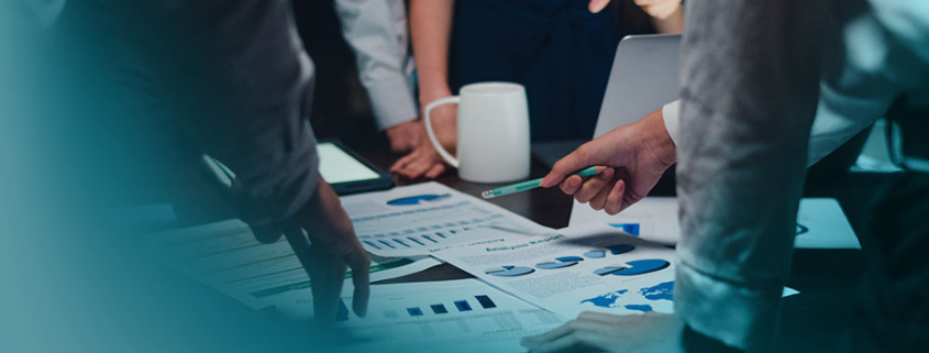 Klassisches Projektmanagement: Dynamische Teambesprechung bei gedämpftem Licht. Detailaufnahme von Händen, die auf Geschäftsdokumente mit Statistiken und Wachstumskurven deuten.