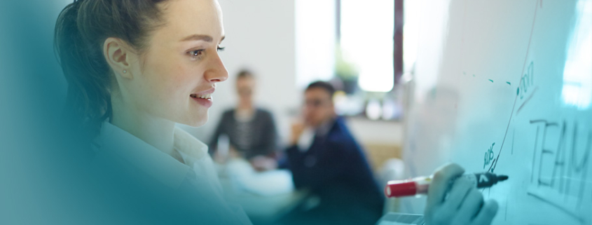 Crossfunktionale Teams: Eine junge Führungskraft in weißer Bluse präsentiert aktiv Ideen am Whiteboard, während im Hintergrund Teammitglieder aufmerksam zuhören.
