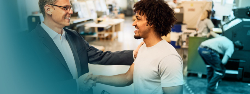 Talentmanagement: Ein Manager in Anzug und ein Mitarbeiter in Arbeitskleidung schütteln sich lächelnd die Hände in einer Werkshalle. Die Szene zeigt Wertschätzung und Anerkennung als wichtigen Bestandteil einer guten Mitarbeiterbindung.