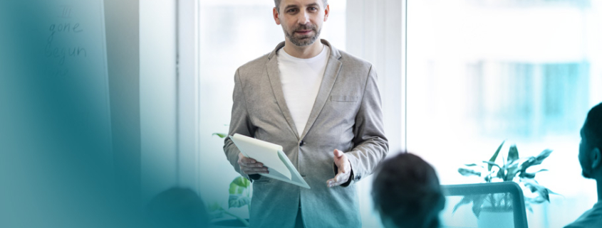 Train the Trainer: Moderne Schulungsatmosphäre mit einem Trainer, der vor einem Whiteboard und hellen Fenstern sein Wissen einer kleinen Teilnehmergruppe vermittelt.