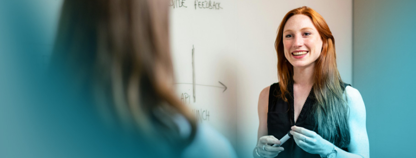Überzeugend präsentieren: Eine kompetente Trainerin in schwarzer Bluse steht vor einem Whiteboard und erklärt mit positiver Ausstrahlung Lerninhalte