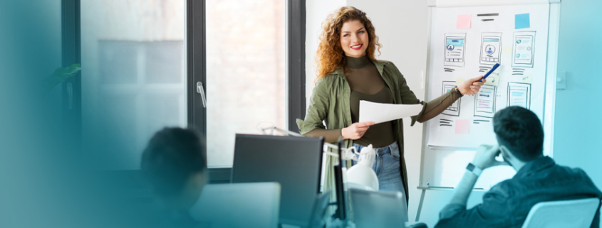 Weiterbildung im Vertrieb: Professionelle Dozentin vermittelt innovative Verkaufsstrategien anhand von UI-Skizzen in moderner Büroumgebung.