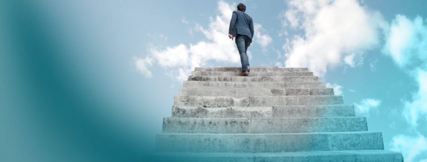 Aufgabe einer Führungskraft: Ein aufstrebender Businessmann auf einer monumentalen Treppe vor blauem Himmel - metaphorisch für Karrierewachstum und Zielsetzung.