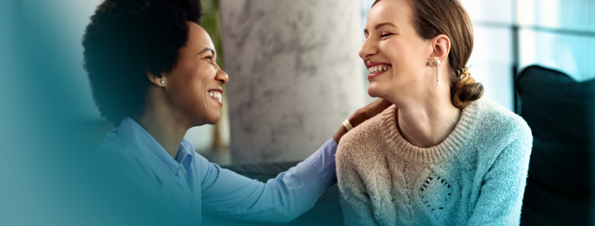 Empathie: Eine Frau legt tröstend ihre Hand auf die Schulter einer anderen Frau, während sie sich lächelnd ansehen. Diese Szene zeigt die Bedeutung von Mitgefühl und Verständnis in zwischenmenschlichen Beziehungen.