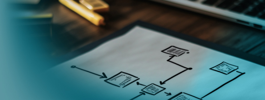 Projektstrukturplan: Handgezeichnetes Flussdiagramm auf weißem Papier mit verbundenen Rechtecken und Pfeilen, im Hintergrund ein Laptop und Büromaterial.