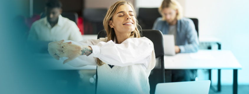 Innere Haltung: Eine entspannte Frau im Büro streckt sich lächelnd am Arbeitsplatz, während Kollegen im Hintergrund arbeiten – Sinnbild für Wohlbefinden und Lernbereitschaft.