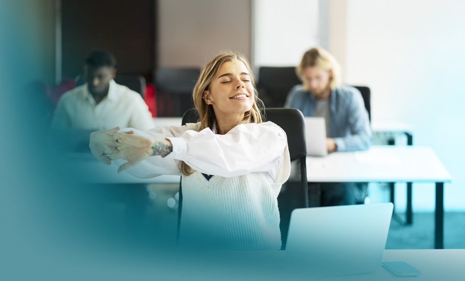 Innere Haltung: Eine entspannte Frau im Büro streckt sich lächelnd am Arbeitsplatz, während Kollegen im Hintergrund arbeiten – Sinnbild für Wohlbefinden und Lernbereitschaft.