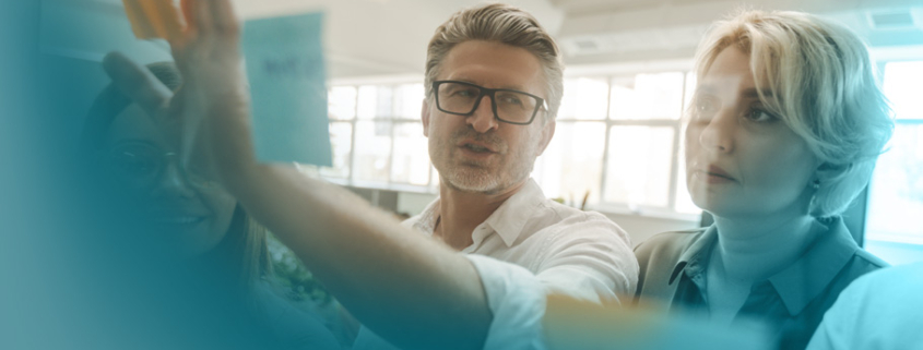 Lernkultur: Drei Fachkräfte teilen Wissen an einer Glaswand mit orangefarbenen Haftnotizen – Visualisierung von Teamlernen in moderner Arbeitsumgebung.