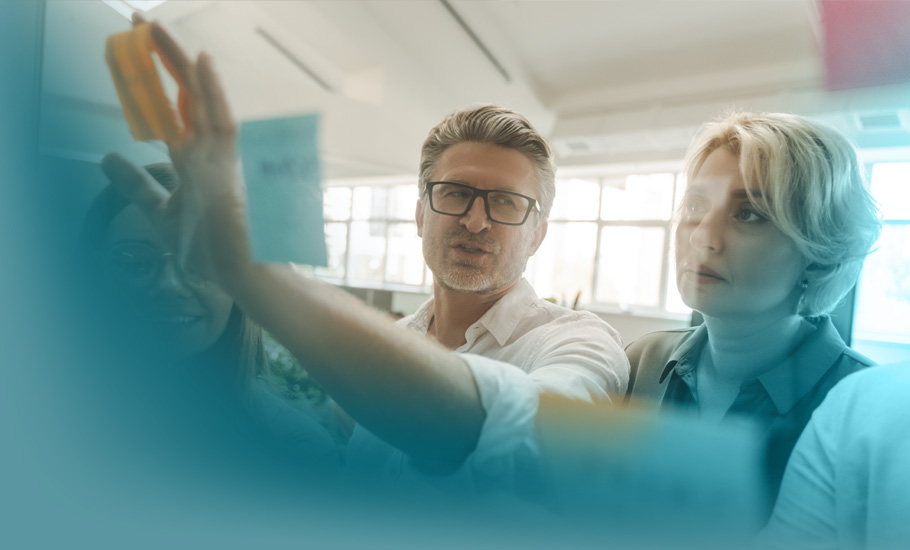 Lernkultur: Drei Fachkräfte teilen Wissen an einer Glaswand mit orangefarbenen Haftnotizen – Visualisierung von Teamlernen in moderner Arbeitsumgebung.