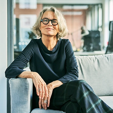 Ältere Frau mit grauen Haaren und runder Brille sitzt entspannt auf hellgrauem Sofa im modernen Büro. Führung 360
