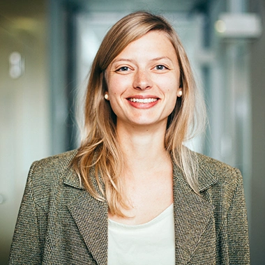 Junge Frau mit blonden Haaren und braunem Blazer lächelt freundlich in die Kamera auf hellem Flur. Führung 360