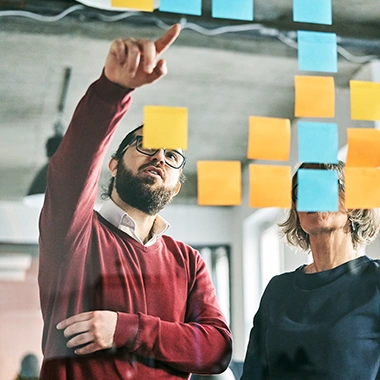 Mann mit Bart und Brille zeigt auf bunte Haftnotizen an Glasscheibe, Frau in Dunkelblau steht daneben. Führung 360