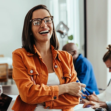 Fröhliche Frau mit Brille und orangefarbener Jacke lacht herzlich, im Hintergrund arbeiten Kollegen in hellem Büro. Strategieentwicklung