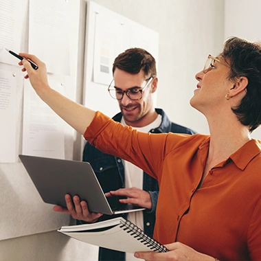 Frau in orangefarbenem Hemd zeigt Mann mit Brille etwas an einem Whiteboard, beide arbeiten konzentriert zusammen. Strategieentwicklung