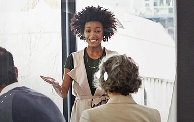 Eine junge, schwarze Frau mit Afro-Frisur präsentiert lächelnd vor zwei Kollegen in einem hellen, modernen Büroraum am Fenster. Change und Organisationsentwicklung