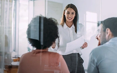 Eine junge Frau in einer weißen Bluse präsentiert lächelnd mit einem Dokument in der Hand vor zwei Kollegen in einem hellen Büro. Change-Management & Leadership