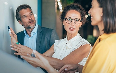 Gespräch am Whiteboard, Frau in Weiß zeigt etwas, Kollege hört zu, unscharfer Vordergrund links, moderne Büroumgebung. Führen mit Zielen