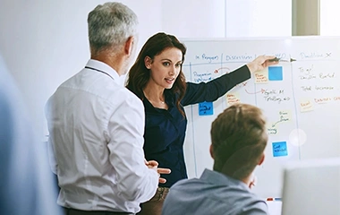 Geschäftsfrau in dunkler Business-Kleidung erklärt enthusiastisch vor Whiteboard mit farbigen Post-its, Kollegen hören zu. Führen von Führungskräften