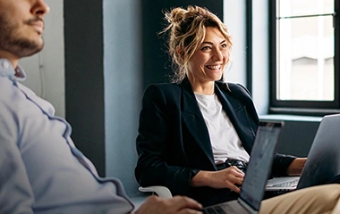 Fröhliche Geschäftsfrau mit Laptop in hellem Büroraum, Team-Kollegen arbeiten im Hintergrund. Grundlagen der Führung