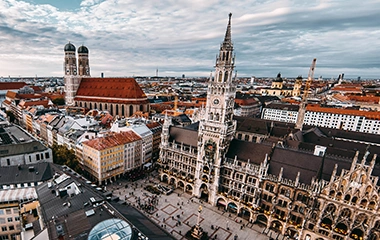 info-und-service-standorte-muenchen München: Luftaufnahme Münchens mit Marienplatz, Frauenkirche und Rathaus bei dramatischem Himmel über der historischen Altstadt mit roten Dächern.