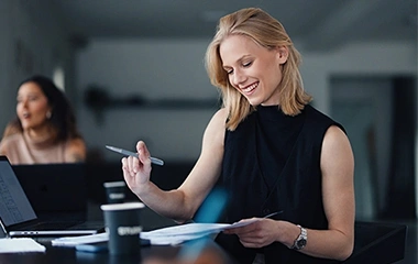 Blonde Frau arbeitet lächelnd mit Stift und Papier am Tisch, Laptop daneben, Kollegin unscharf im Hintergrund. Agiles Projektmanagement