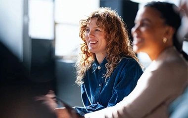 Frau mit Locken lacht in einer Gruppe, helle Büroumgebung mit weichem Licht und Sitzreihe. Grundlagen Projektmanagement