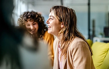 Zwei Frauen sitzen auf einem Sofa und lachen, unscharfe Person im Vordergrund, helles Büro mit Glaswänden. Hybrides Projektmanagement