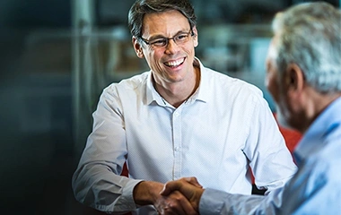 Handschlag zwischen zwei Männern am Tisch, Mann mit Brille lächelt, moderne Umgebung mit Glas und neutralen Farben. Strategische Kommunikation