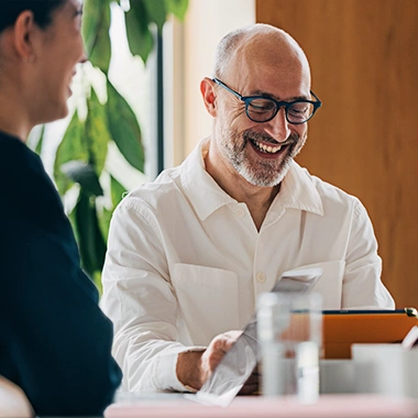 Lächelnder Mann mit Brille und Bart im weißen Hemd sitzt am Tisch und schaut auf Tablet, daneben Person von hinten. Strategieentwicklung