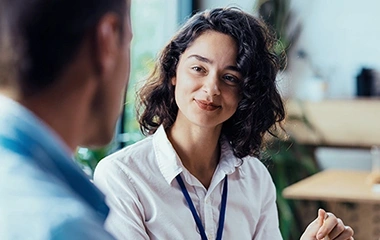 Junge Frau mit dunklen Haaren und weißer Bluse hört aufmerksam einem Mann zu, weiche Bürohintergründe mit Pflanzen und Fenstern. Key Account Management