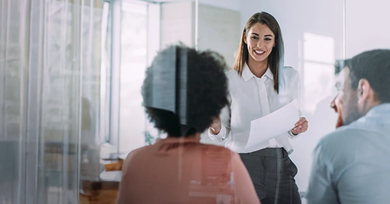 Eine junge Frau in einer weißen Bluse präsentiert lächelnd mit einem Dokument in der Hand vor zwei Kollegen in einem hellen Büro. Change-Management & Leadership